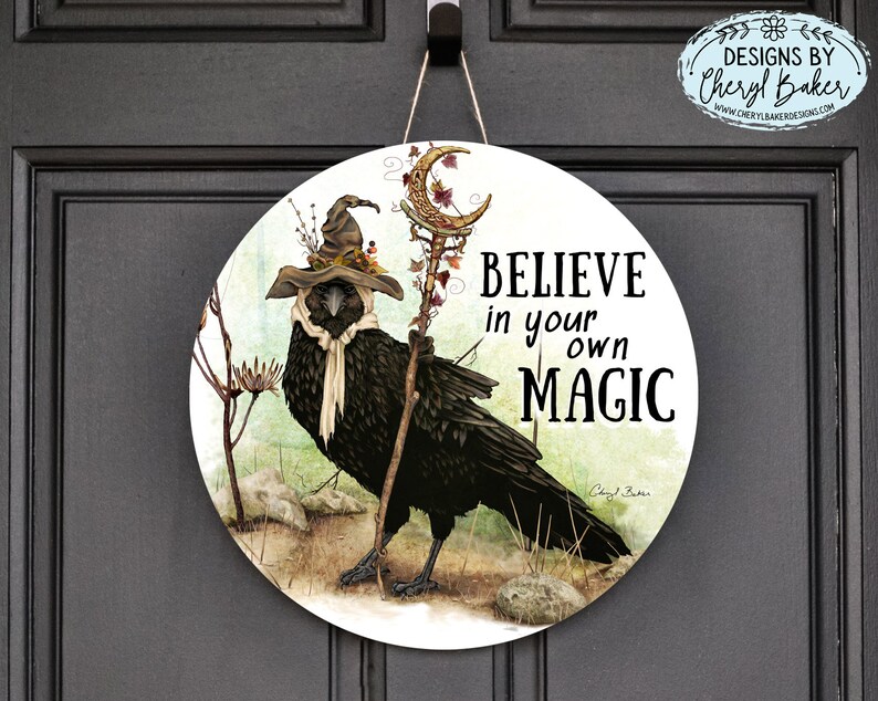 Puede incluir: Un letrero redondo de madera con un cuervo negro que lleva un sombrero de bruja y sostiene un bast&oacute;n. El letrero tiene un fondo verde y marr&oacute;n con el texto "Cree en tu propia magia" en letras negras.
