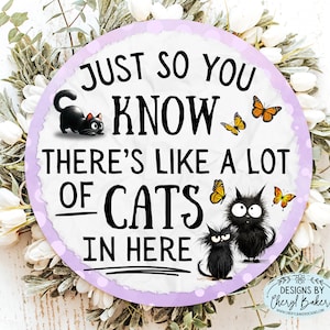 May include: Round sign with the text "JUST SO YOU KNOW THERE'S LIKE A LOT OF CATS IN HERE." The sign has a purple border and features illustrations of black cats and yellow butterflies. The sign is surrounded by a white floral wreath.