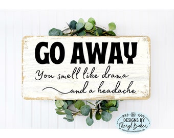 Könnte beinhalten: Ein weißes Holzschild mit schwarzem Text, der "GO AWAY You smell like drama and a headache" lautet. Das Schild ist abgenutzt und hat einen rustikalen Look. Das Schild ist von Grün umgeben.