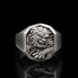 Puede incluir: Un anillo de plata con una cara octogonal que presenta un diseño detallado en tres dimensiones de una criatura mítica. El anillo está colocado sobre un fondo negro.