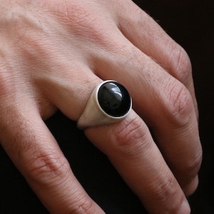 May include: A silver ring with a large, round, black stone. The ring is set in a simple, classic style. The black stone is polished and reflects light. The ring is on a finger.