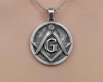 May include: Silver-toned pendant necklace featuring a circular pendant with a Masonic symbol. The pendant displays a compass, square, and the letter "G" in the center. The necklace has a delicate chain and a simple clasp.