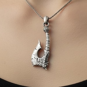 May include: A silver-colored pendant necklace featuring a stylized hook design. The pendant has intricate detailing, including textured surfaces and decorative elements. The necklace chain is also silver and appears to be a woven style.