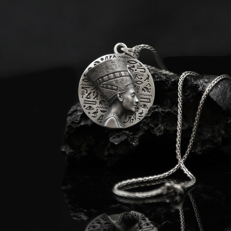 May include: Silver pendant necklace featuring a detailed profile of an Egyptian queen wearing a headdress with hieroglyphics. The pendant is round and hangs from a silver chain.