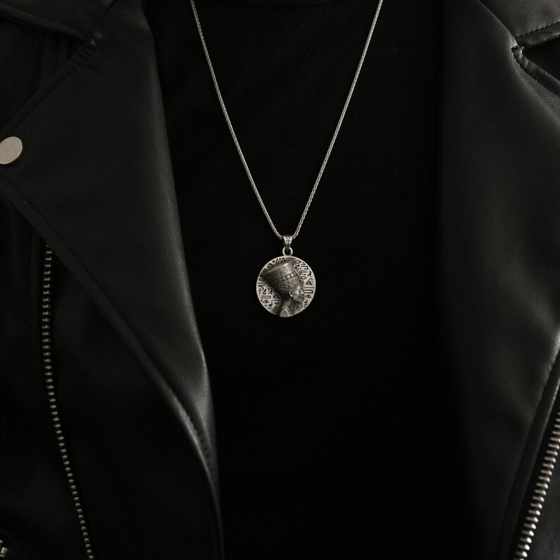 May include: A silver chain necklace with a round pendant featuring a detailed profile of an Egyptian queen. The pendant is surrounded by hieroglyphs.