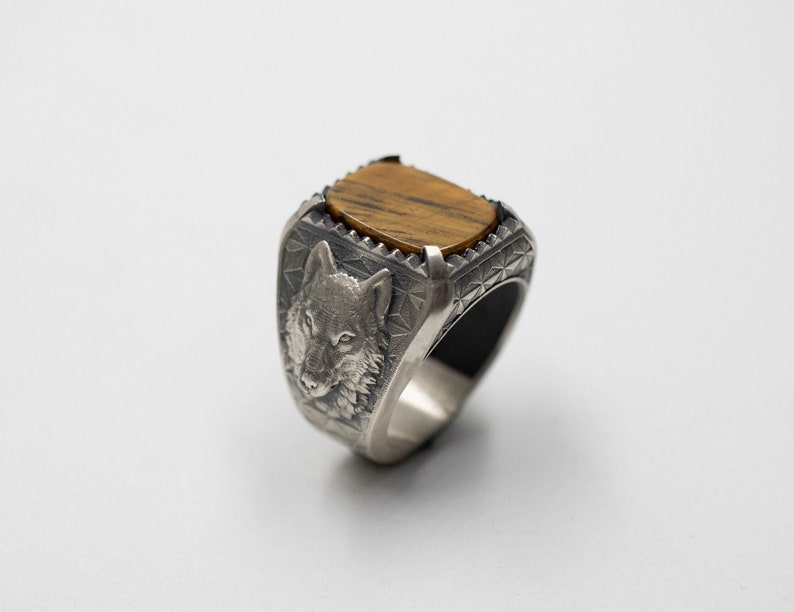 Puede incluir: Un anillo de plata con un dise&ntilde;o de cabeza de lobo y una piedra de ojo de tigre marr&oacute;n.