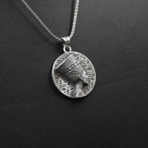 May include: Silver necklace with a round pendant featuring a detailed profile of an Egyptian queen wearing a headdress with hieroglyphics.