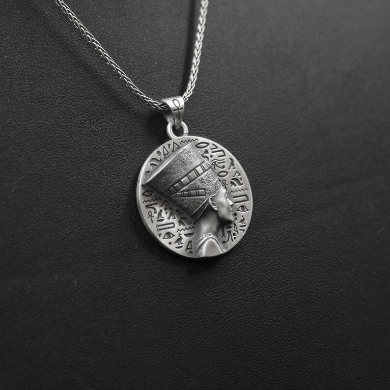 May include: Silver necklace with a round pendant featuring a detailed profile of an Egyptian queen wearing a headdress with hieroglyphics.
