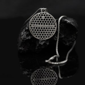 May include: A silver necklace with a round pendant featuring a honeycomb pattern and the word "Personalization" engraved on the back. The pendant is hanging on a black rock.