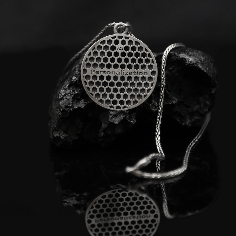 May include: A silver necklace with a round pendant featuring a honeycomb pattern and the word "Personalization" engraved on the back. The pendant is hanging on a black rock.