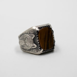 Puede incluir: Un anillo de plata con un dise&ntilde;o de cabeza de lobo y una piedra de ojo de tigre marr&oacute;n.