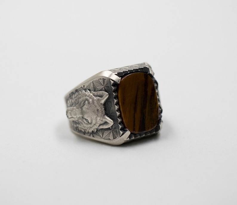 Puede incluir: Un anillo de plata con un dise&ntilde;o de cabeza de lobo y una piedra de ojo de tigre marr&oacute;n.