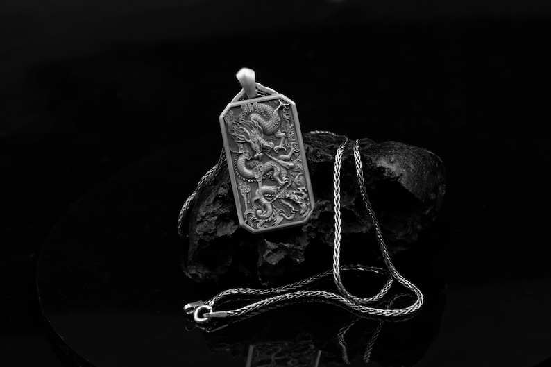 May include: A silver pendant necklace featuring a detailed dragon design. The pendant is rectangular and has a textured surface. The chain is silver and has a box link style.