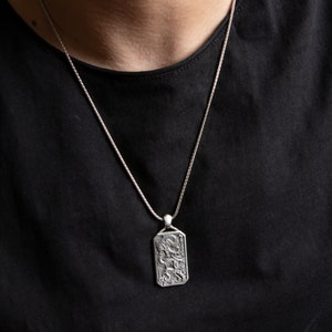 May include: A silver chain necklace with a rectangular pendant featuring an intricate design. The pendant has a detailed, textured surface.