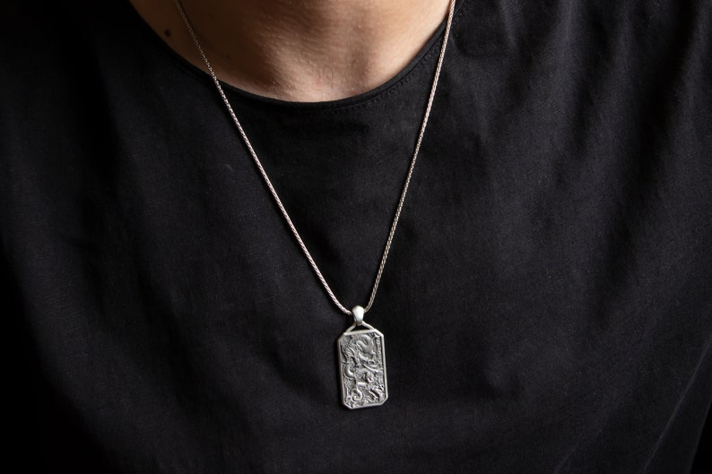 May include: A silver chain necklace with a rectangular pendant featuring an intricate design. The pendant has a detailed, textured surface.