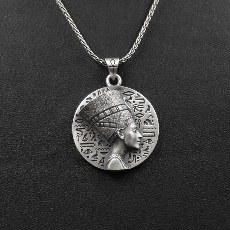 May include: Silver pendant necklace featuring a detailed profile of an Egyptian queen wearing a headdress, surrounded by hieroglyphs.