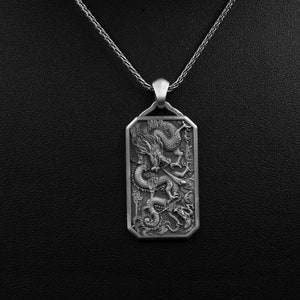 May include: A silver pendant necklace featuring a detailed dragon design. The pendant is rectangular and has a textured surface.