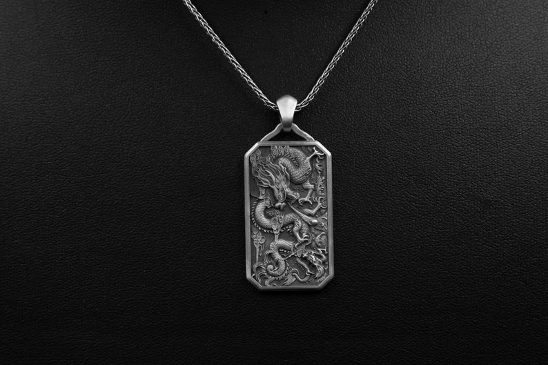 May include: A silver pendant necklace featuring a detailed dragon design. The pendant is rectangular and has a textured surface.