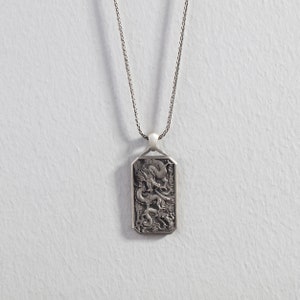 May include: A silver necklace with a rectangular pendant featuring an intricate, detailed design of a dragon. The pendant has a slightly rounded top and bottom.