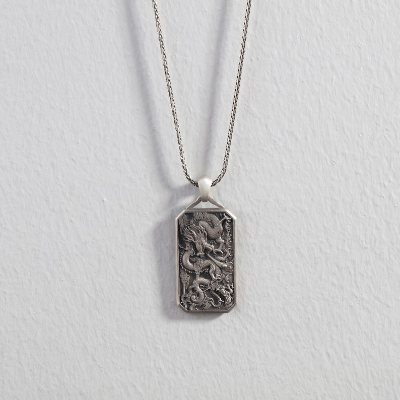 May include: A silver necklace with a rectangular pendant featuring an intricate, detailed design of a dragon. The pendant has a slightly rounded top and bottom.