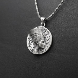 May include: Silver necklace with a round pendant featuring a detailed profile of an Egyptian queen wearing a headdress. The pendant is surrounded by hieroglyphs.