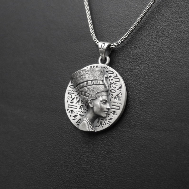 May include: Silver necklace with a round pendant featuring a detailed profile of an Egyptian queen wearing a headdress. The pendant is surrounded by hieroglyphs.
