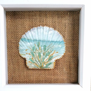 May include: A framed artwork featuring a painted seashell. The shell is decorated with a light blue ocean scene and green and gold plants. The background has a burlap texture.