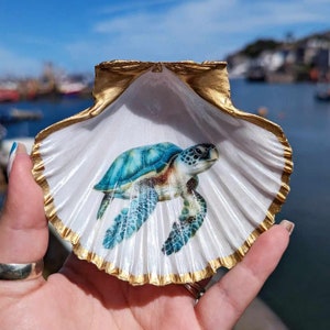 May include: A white seashell with a gold trim, painted with a blue and green sea turtle. The shell is held in a hand.