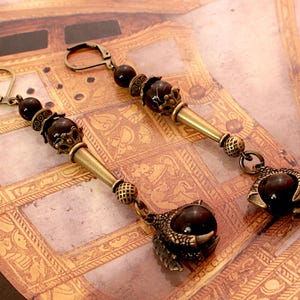 May include: A pair of dangle earrings with brown and gold accents. Each earring features a large, dark brown gemstone surrounded by a gold-toned, ornate design. The earrings are suspended from a gold-toned chain with smaller brown beads.