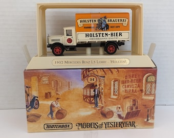 May include: A white Matchbox Models of Yesteryear die-cast toy truck with a Holsten-Bier logo on the side. The truck is in a clear plastic display case with a brown cardboard box. The box has a picture of a German-style building with a sign that says "BIERE, MER-BEER, CERVEZA, BIRRA".