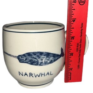 May include: White ceramic mug with a blue and white narwhal illustration and the word "NARWHAL" printed on the front. The mug has a handle on the right side.