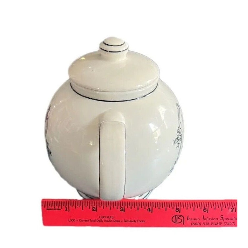 May include: White ceramic teapot with a lid and handle, featuring black trim and decorative illustrations. A red ruler with text is at the bottom. The teapot is designed for brewing tea.
