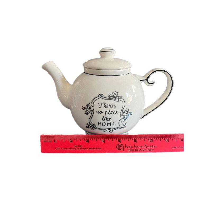 May include: Cream-colored ceramic teapot with a lid and curved handle. The teapot has black trim and a decorative frame with the text "There's no place like HOME". A red ruler is at the base of the teapot.
