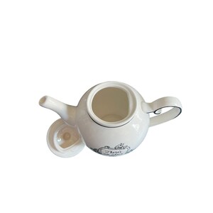 May include: White ceramic teapot with a curved handle and spout. The teapot has a black trim and a lid. The teapot has the text "There's no place like home" in a decorative frame.
