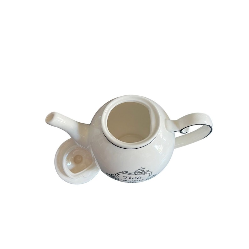 May include: White ceramic teapot with a curved handle and spout. The teapot has a black trim and a lid. The teapot has the text "There's no place like home" in a decorative frame.