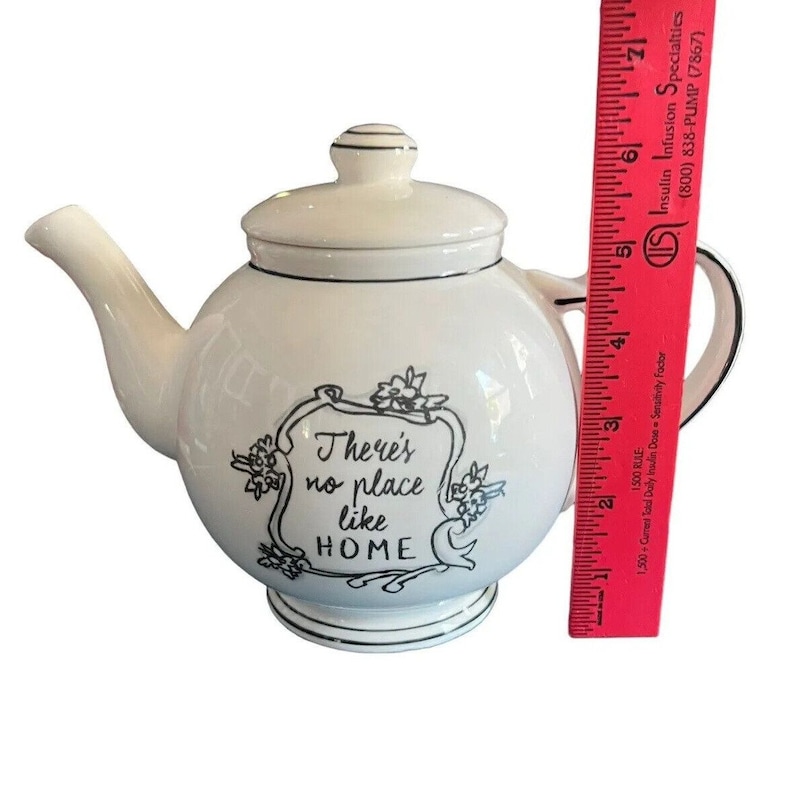 May include: White ceramic teapot with a lid and spout, featuring black trim and the phrase "There's no place like HOME." The teapot is approximately 5 inches tall. A decorative item for the kitchen or dining room.