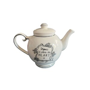 May include: Cream-colored ceramic teapot with a lid and curved handle. The teapot features a black outlined design with the phrase "Home is where the HEART is" in a decorative frame. The spout and base are also outlined in black.