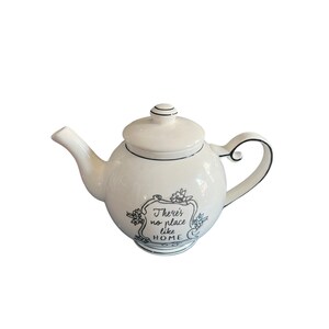 May include: A white ceramic teapot with a rounded body, spout, and handle. The lid has a small knob. The teapot is decorated with black trim and a black and white floral design with the text "There's no place like HOME".