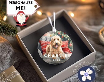 May include: A round ceramic ornament featuring a watercolor painting of a dog sitting on a red and white patterned pillow. The dog is a light brown color with a white chest and a black nose. The ornament is personalized with the name "CHARLIE" and the year "2024".