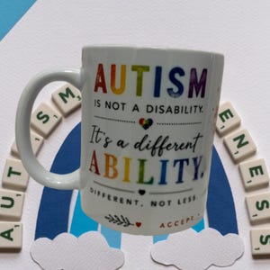 May include: A white ceramic mug featuring the rainbow-colored text AUTISM IS NOT A DISABILITY. IT'S A DIFFERENT ABILITY. DIFFERENT, NOT LESS. ACCEPT. The design includes a small puzzle piece heart, a leaf sprig, and a small red heart icon.