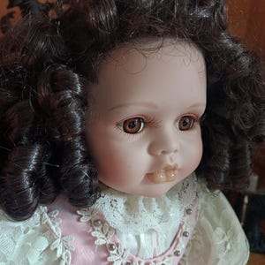 May include: A porcelain doll with brown, curly hair and brown eyes. The doll is dressed in a pink dress with white lace trim and pearl accents. The doll's face has a realistic appearance with painted features.