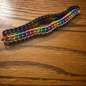 May include: Two chainmail bracelets. One is a rainbow of colors, and the other is a darker rainbow. The bracelets are made of small, interlocking metal rings. The bracelets are laying on a wooden surface.