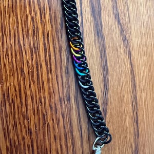 Puede incluir: Correa de muñeca de cota de malla negra con un acento arcoíris. La cota de malla está hecha de pequeños anillos entrelazados. En la parte inferior hay un cierre de mosquetón plateado. La correa se muestra sobre una superficie de madera.