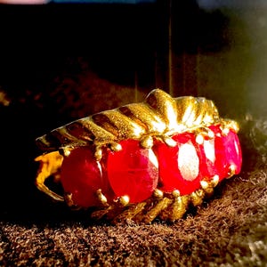 May include: A gold ring featuring a row of five round, vibrant red gemstones set horizontally. The metal band displays a textured, ridged design along the top edge, with each stone secured by small, rounded prongs in a classic, structured arrangement.
