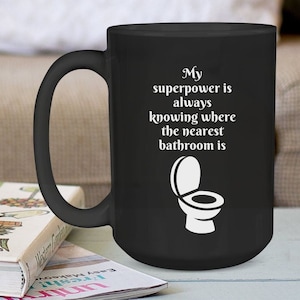 May include: Black coffee mug with a white design that says "My superpower is always knowing where the nearest bathroom is" and a white silhouette of a toilet.