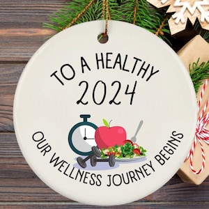 May include: White ceramic ornament with the text "To a Healthy 2024 Our Wellness Journey Begins" and a graphic of a stopwatch, apple, dumbbells, and a plate of salad.