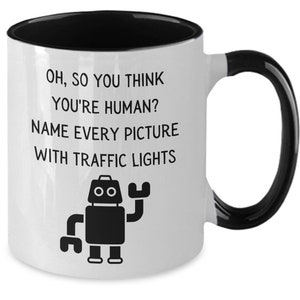 May include: White coffee mug with a black rim and handle. The mug has a black robot illustration and text that reads "Oh, so you think you're human? Name every picture with traffic lights."