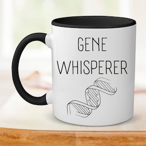 May include: White ceramic coffee mug with a black rim and handle. The mug has a black and white illustration of a DNA strand and the text "GENE WHISPERER" printed on it.