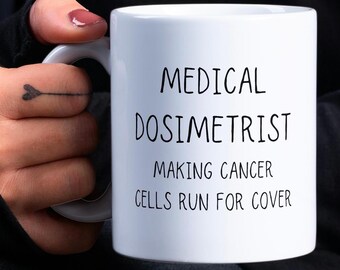 Könnte beinhalten: Weißer Keramikbecher mit dem Text "MEDICAL DOSIMETRIST MAKING CANCER CELLS RUN FOR COVER" in Schwarz. Der Becher wird von einer Person gehalten.