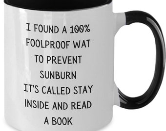 May include: White ceramic mug with a black interior, handle, and rim. The mug features the text: "I FOUND A 100% FOOLPROOF WAT TO PREVENT SUNBURN IT'S CALLED STAY INSIDE AND READ A BOOK" in black font.